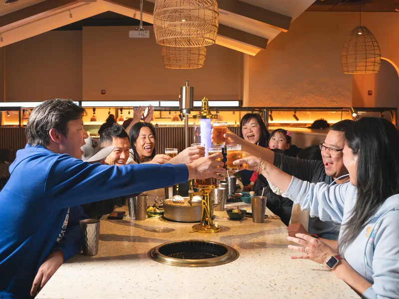Family and friends clink beers around a lit beer tower at a long grill table under rattan lamps