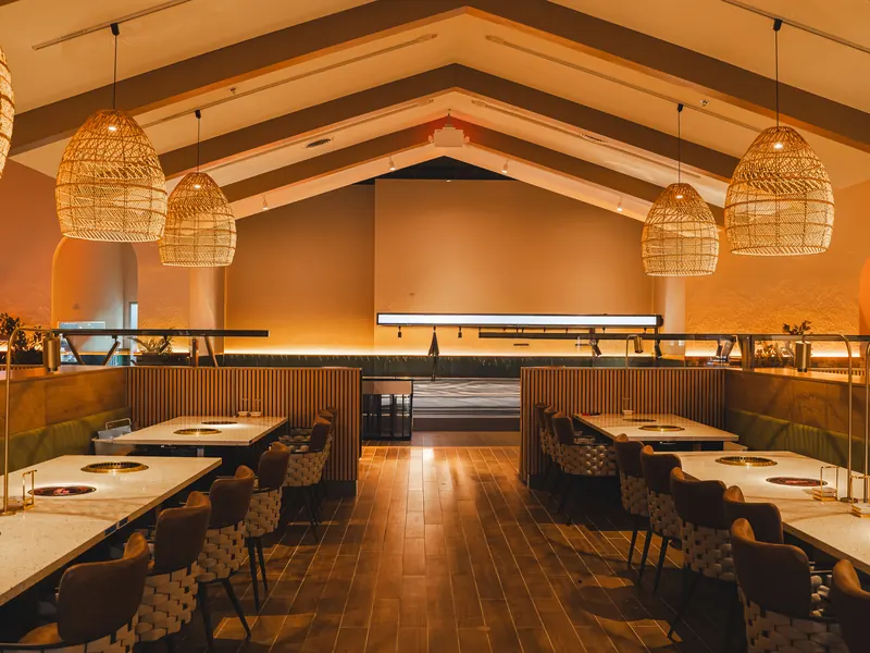 The main dining hall under a vaulted beamed ceiling, rattan pendant lamps glowing over rows of grill tables