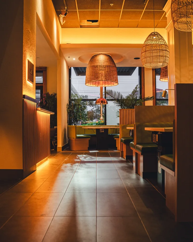 The entry walkway at dusk: warm-lit wood panels lead past green booths to a window table under a rattan lamp and copper grill hood