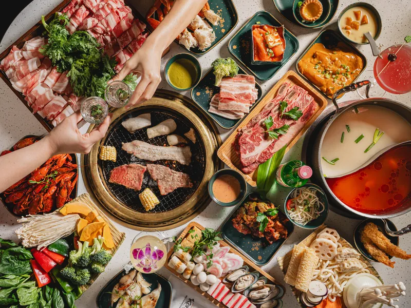 Overhead view of a full Kanpai table: a charcoal grill of short rib and corn, a split hotpot of white and red broth, platters of sliced pork and beef, crawfish, seafood and a toast of soju glasses