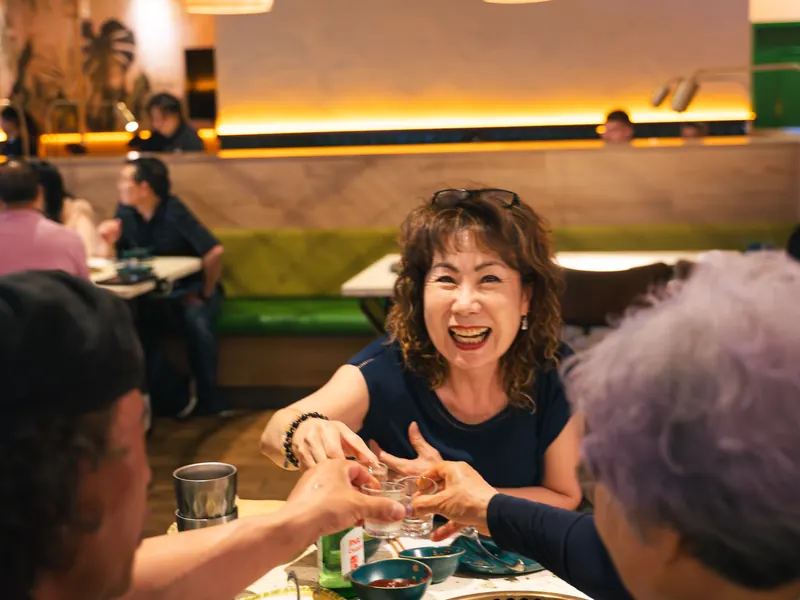 A laughing guest clinks soju glasses with friends at a grill table, green booths behind